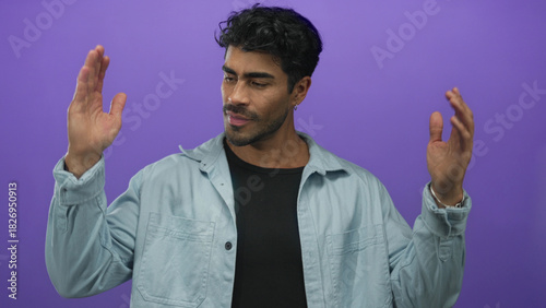 Young hispanic man in a light blue denim shirt holding hands apart measuring width in a purple studio; size evaluation.