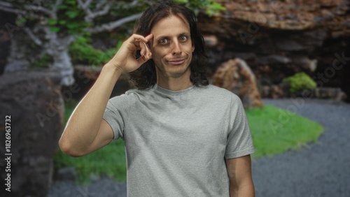 Young hispanic man with finger to temple and long hair wearing gray t shirt in forest path with green foliage; introspection.