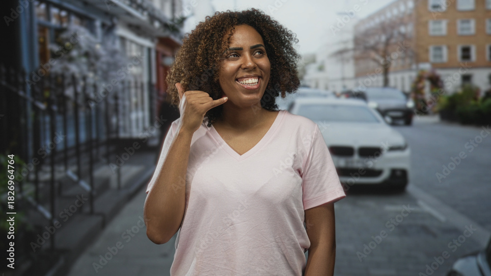 Fototapeta premium Young woman smiling with hand to ear and pinky to mouth call gesture on a city street with parked car; joy connection.
