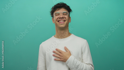 Fotografie Young hispanic man wearing glasses with hand on chest laughing in studio setting; amusement gratitude