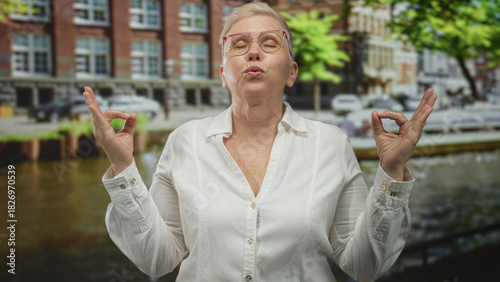 Foto Woman making ok sign with both hands, eyes closed and puckered lips, glasses visible beside a canal on amsterdam street with brick buildings; mindfulness calm
