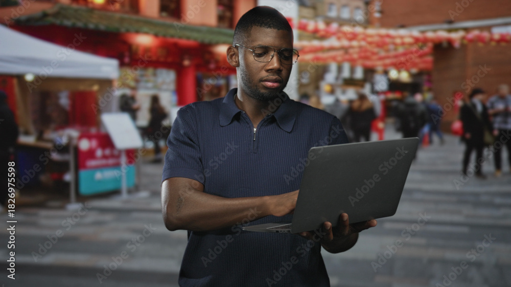 Fototapeta premium Young black man wearing glasses typing on laptop, hands visible in busy street market; concentration productivity focus.