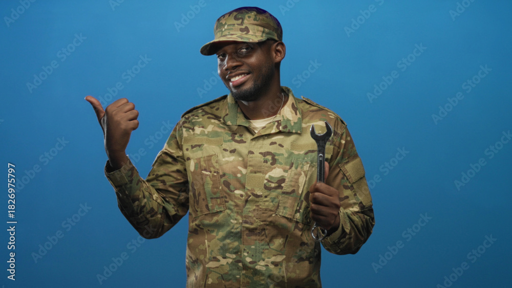 Fototapeta premium Man in camouflage uniform and cap holding wrench and pointing thumb to left in studio; confidence teamwork.