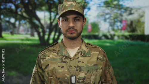 Man wearing green camouflage uniform, cap, and dogtags looks at camera while standing in a forest; duty.