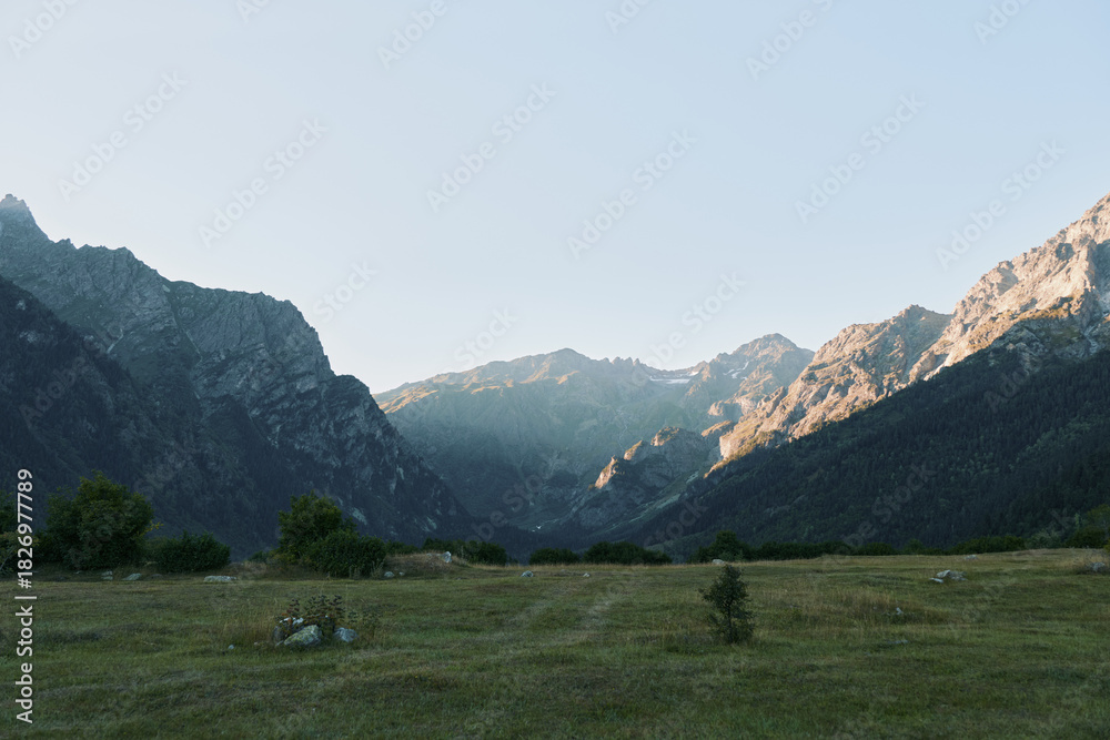 Fototapeta premium A broad mountain landscape unfolds over a grassy valley under a clear sky. Rugged slopes frame the scene, inviting reflection and exploration in a peaceful outdoor setting.