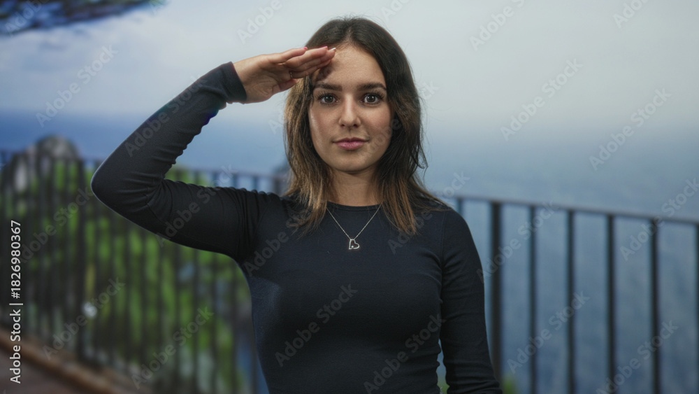 Obraz premium Young woman salutes with hand to forehead on building terrace by railing overlooking ocean view; confidence.