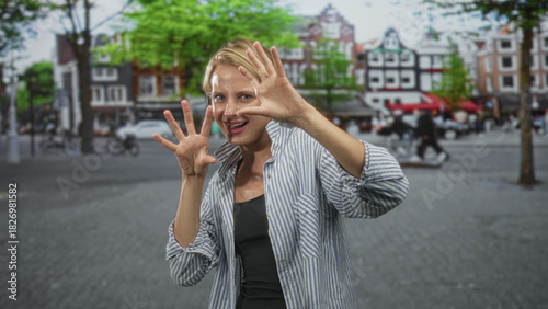 Fotografie Blond woman in striped shirt shows open palms, spreads fingers and pushes hands toward camera on an urban street framed by canalside houses and trees; playful curiosity