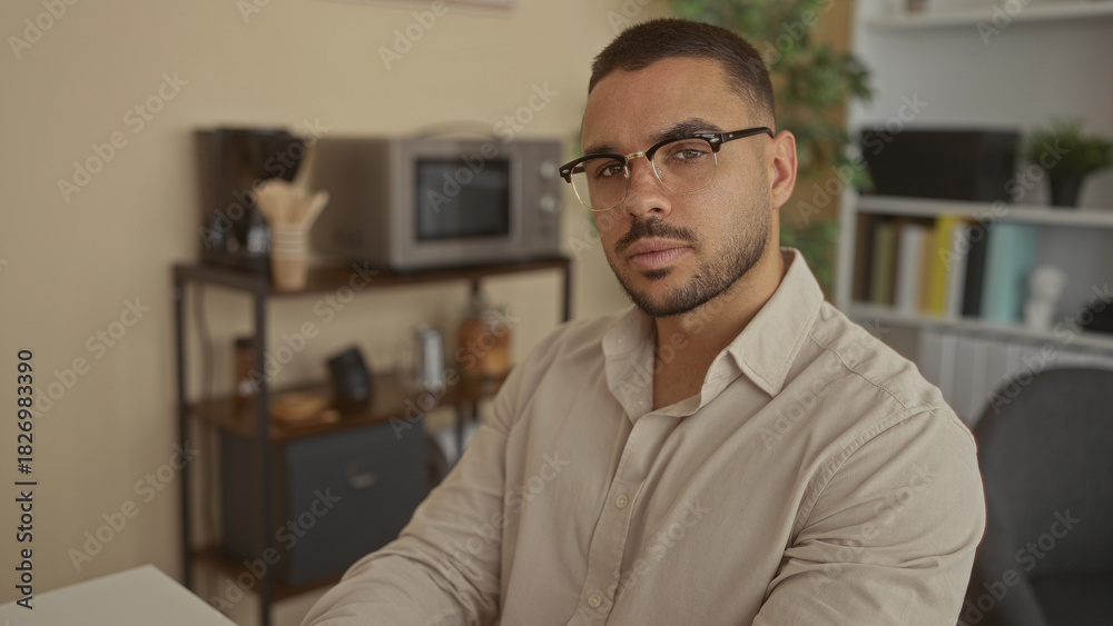 Fototapeta premium Young hispanic man wearing glasses glances to the side at a coffee station on a shelf in an office workspace; contemplation.