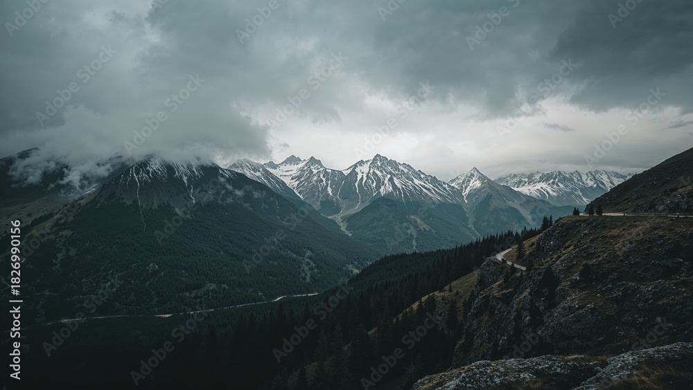 Fototapeta premium Snow-capped mountains under stormy clouds with dense forests and rugged terrain. Mountain landscape, wilderness, scenic nature view.
