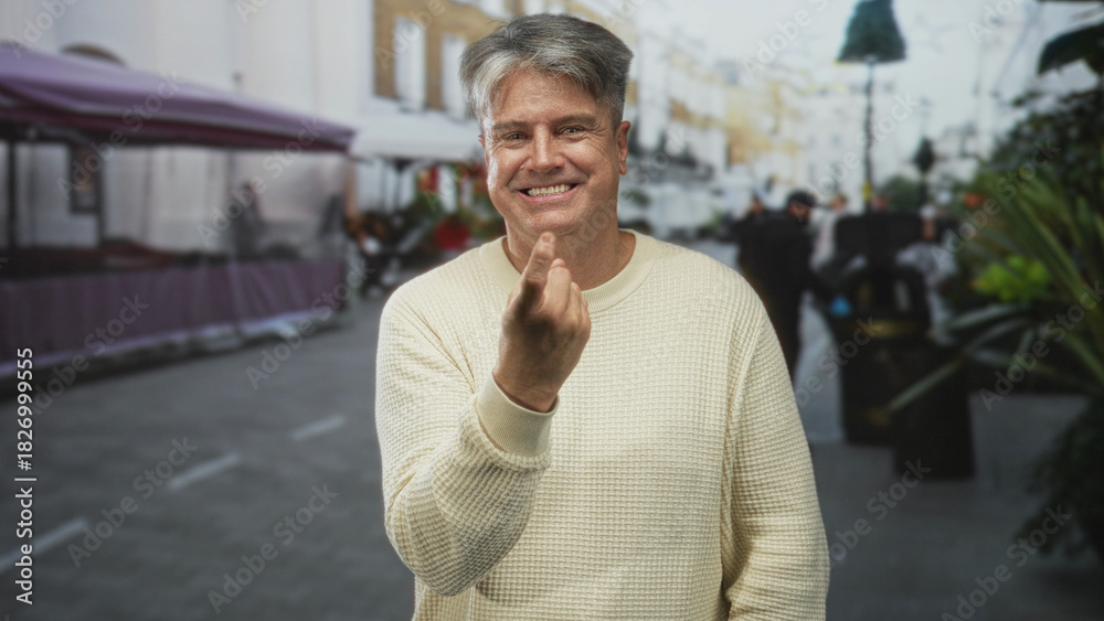Naklejka premium Senior man smiling, beckoning with finger on a street market beside stalls and planters; playful invitation.
