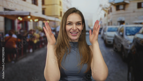 Woman frames face with hands while smiling on busy city street lined with parked cars and sidewalk cafes; playful joy. Free Download Woman frames face with hands while smiling on busy city street lined with parked cars and sidewalk cafes; playful joy. - GFXLoad