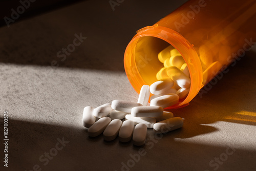 A dramatic, low-key photograph of capsules and pills spilling from an orange bottle onto a concrete surface, highlighted by hard light and deep shadow.