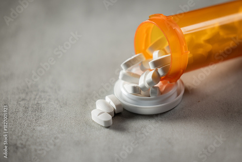 A dramatic, low-key photograph of capsules and pills spilling from an orange bottle onto a concrete surface, highlighted by hard light and deep shadow.