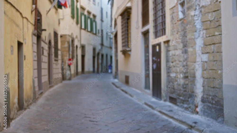 Fototapeta premium Narrow cobblestone alley defocused bokeh with soft focus on textured stone and stucco facades, receding urban passage and muted warm tones; background backplate copyspace calm.