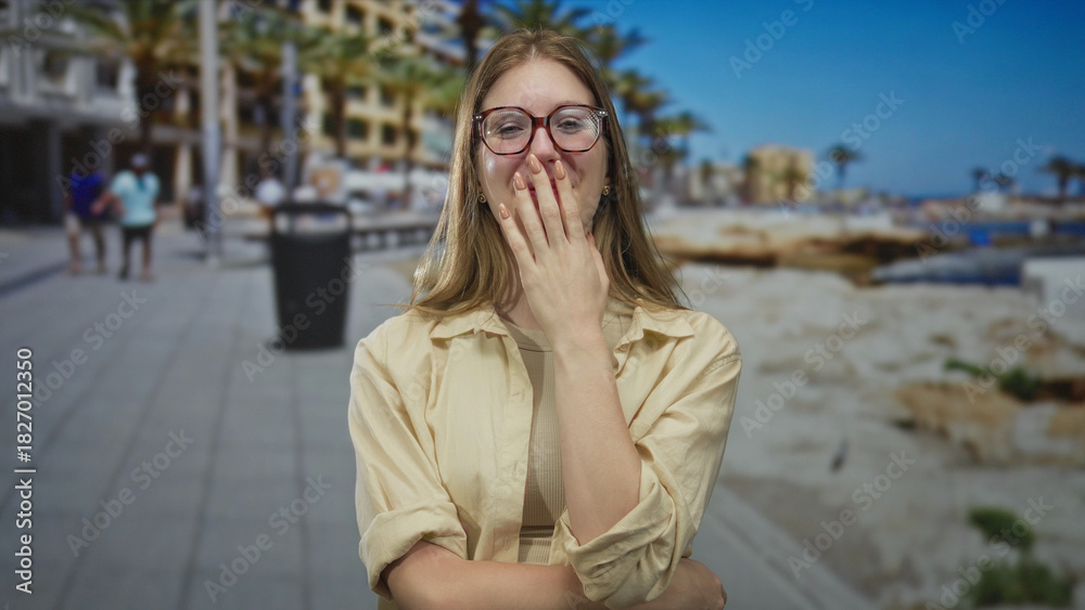 Fototapeta premium Young blonde woman wearing glasses laughs and covers mouth with hand on sunlit street by building; joy.