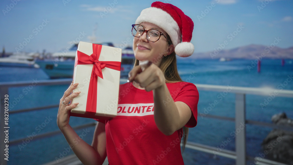 Obraz premium Woman volunteer in red santa hat smiling holds gift box and points finger on street; holiday joy and giving spirit.