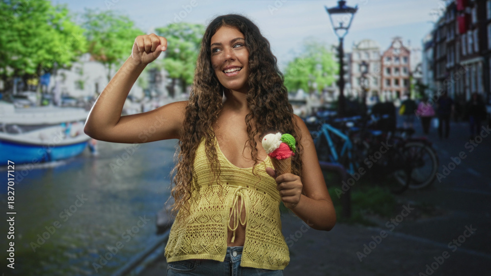 Fototapeta premium Woman holding ice cream cone and raising fist on street by a canal, smiling in yellow knit top; joyful carefree summer.
