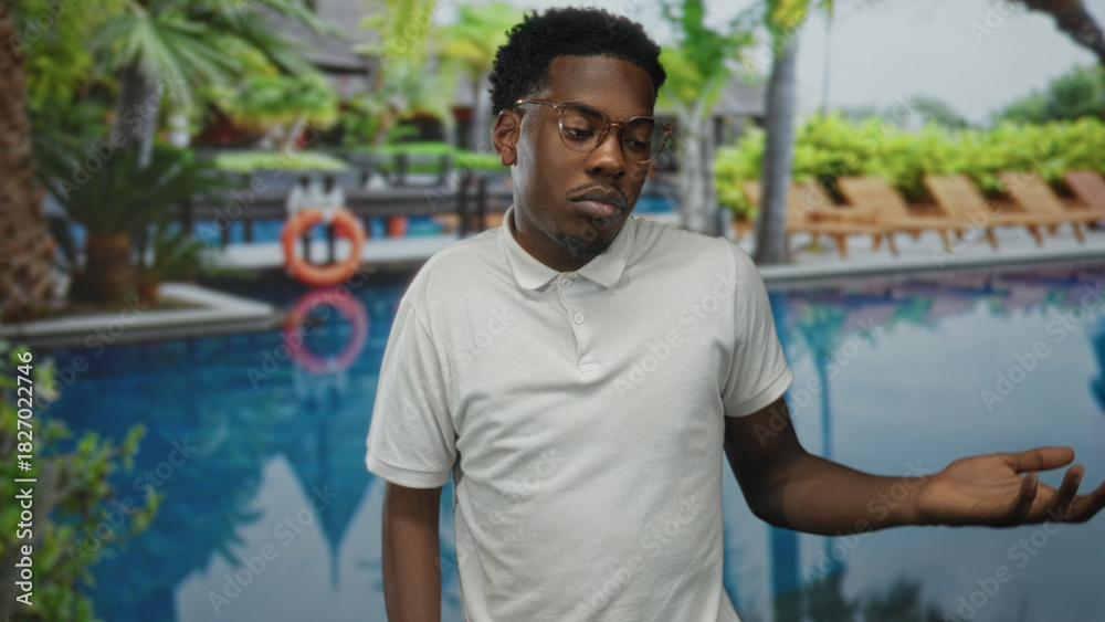 Fototapeta premium Young african american man casually shrugs at pool outside a resort building; uncertainty reflection.