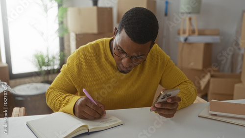 Man writing notes with smartphone in studio stacked with moving boxes and notebooks; concentration productivity.