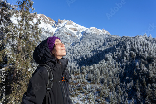 A person enjoys the warmth of the sun while standing in a  snowy mountain landscape. With eyes closed and a peaceful smile, they embrace the crisp air and stunning alpine scenery 