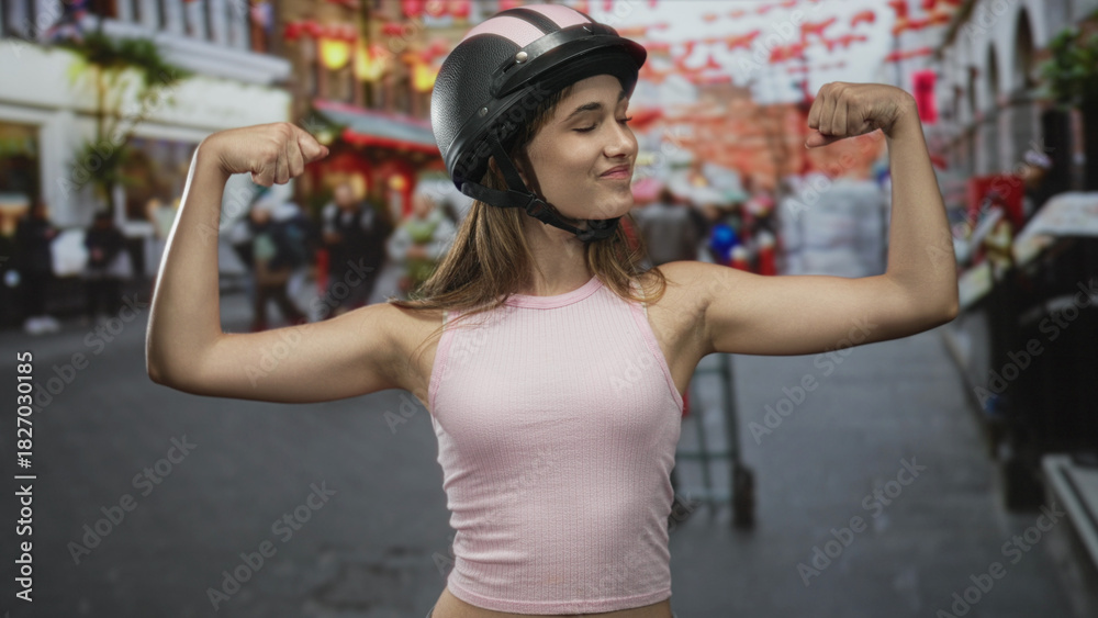 Fototapeta premium Young woman in pink tank top and helmet flexes biceps while smiling on a busy street market; confidence empowerment.