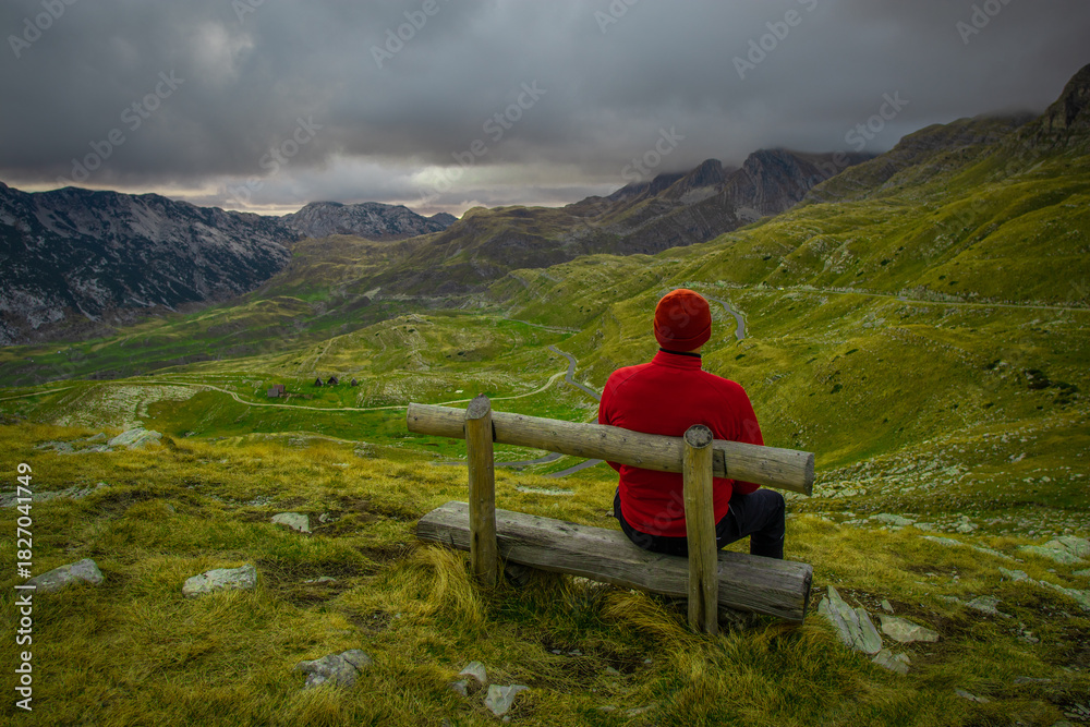 Naklejka premium A person in a red jacket sits on a wooden bench overlooking a dramatic mountain landscape under dark, moody clouds.