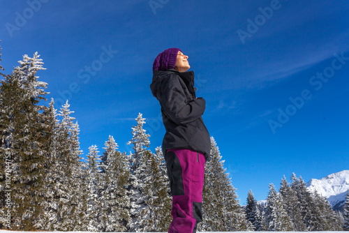 A person enjoys the warmth of the sun while standing in a  snowy mountain landscape. With eyes closed and a peaceful smile, they embrace the crisp air and stunning alpine scenery 