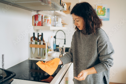 Woman with dark hair wearing a cozy gray sweater is cleaning a modern kitchen countertop with an orange cloth, showcasing a tidy home environment and domestic care