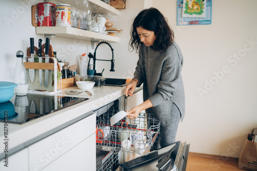Valokuva Asian woman in gray sweater unloading clean dishes from a modern dishwasher in a