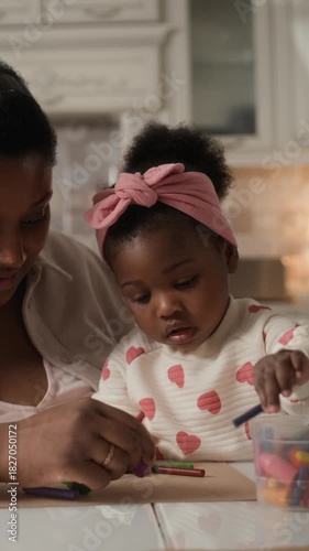 Vertical of young Black mom teaching her toddler daughter how to draw on paper with crayons and kissing her