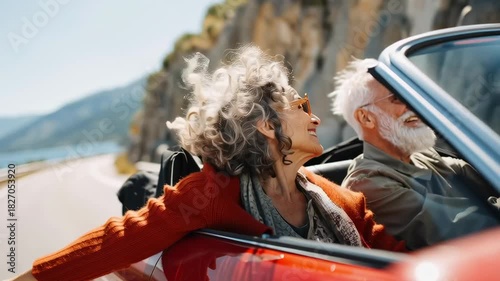 A couple of older people are driving a red convertible car and waving. Scene is happy and carefree