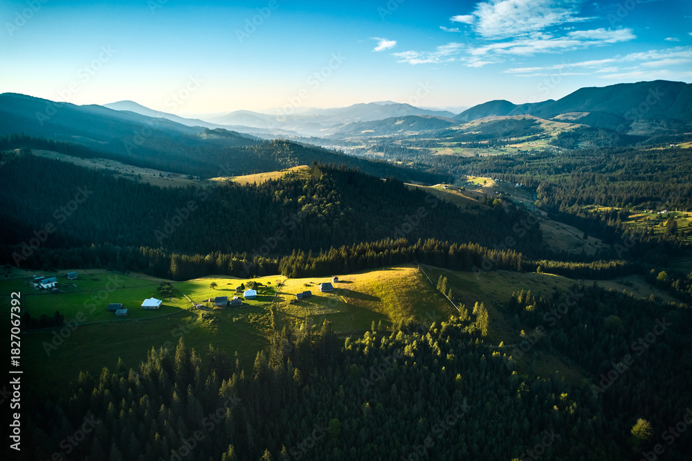 Obraz premium Aerial view of picturesque valley with rolling hills, lush forests, and scattered houses. Sunlight bathes landscape in warm glow, highlighting vibrant greens and creating serene atmosphere.