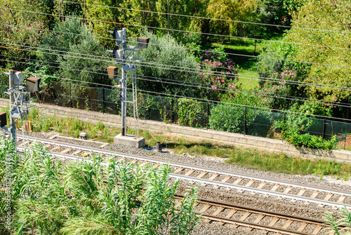 Railway tracks surrounded by greenery, overhead power lines, and signal lights on a sunny day.