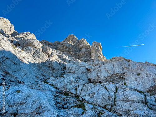 A rugged limestone rock face rises steeply towards the deep blue sky, marked by a faint airplane trail, capturing raw beauty of the Julian Alps near Jof di Montasio in Friuli Venezia Giulia, Italy.