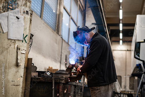 Worker welding metal parts at a workshop table. Bright sparks and industrial atmosphere showcasing precision metal fabrication, craftsmanship, and professional welding work.