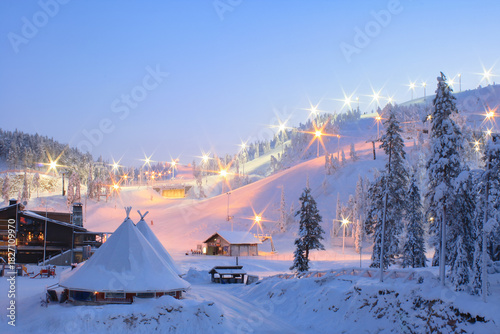 Picturesque winter sunrise at Ruka Ski Resort in Finland, with snow-covered slopes, glowing lights, frosted trees and magical morning atmosphere in a Nordic landscape.