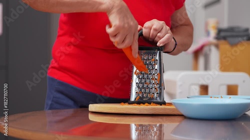 A person is grating fresh carrots in a well-lit kitchen. The kitchen is warm and inviting