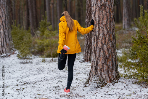 Sporty woman holding a pine tree while stretching in a snow-covered forest, concept of cold weather workout and outdoor fitness routine.