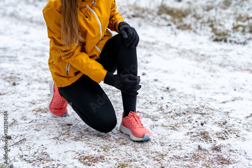 Female athlete forced to stop during a winter run, bending down and clutching her lower leg and knee. Image represents overuse injury, shin splints, and the risks of training in cold conditions.