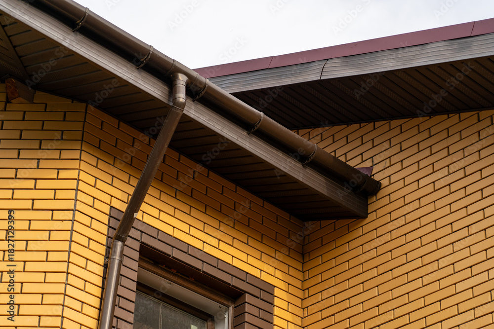 Obraz premium Close-up view of house corner featuring textured yellow brick wall, complete with brown metal gutter system, soffit, and fascia against overcast sky