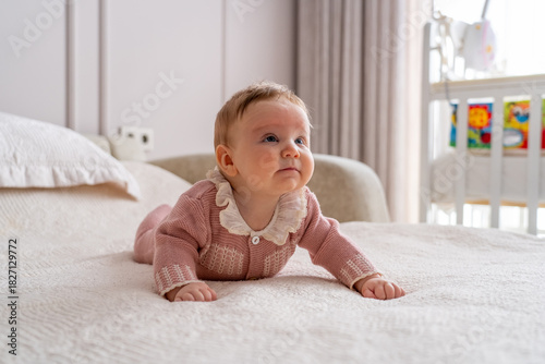 Sweet baby girl in charming pink knitted outfit with ruffled collar practicing tummy time on soft bed, gazing up in bright nursery