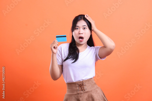 A young woman looks shocked and confused while holding a blue credit card, possibly due to a financial surprise or mistake, isolated on orange background