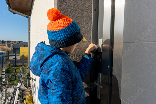 Boy in blue winter jacket and knitted hat with pompom unlocking front door with key on sunny day. Concept of childhood independence, returning home from school, responsibility and daily routine life. 