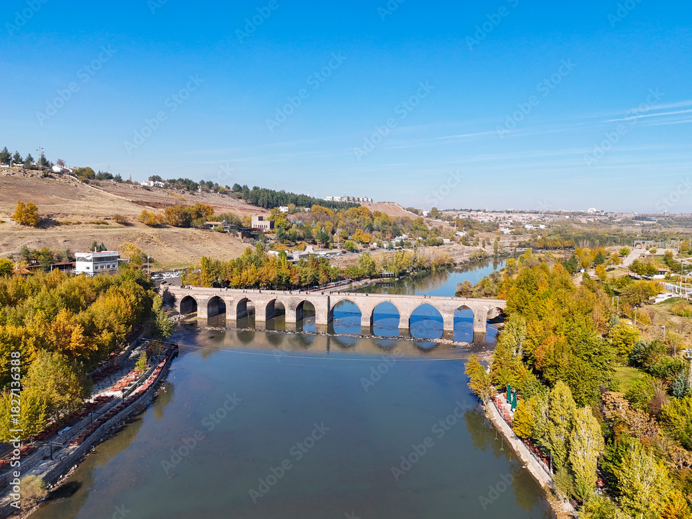 Fototapeta premium Stunning aerial view of the historic Ten-Eyed Bridge (On Gözlü Köprü) over the Tigris River. The stone arches and their reflection are framed by vibrant autumn trees under a bright blue sky.