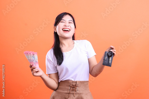 A cheerful woman is holding a car key and a fan of money, expressing excitement and satisfaction with her new purchase isolated on orange background.