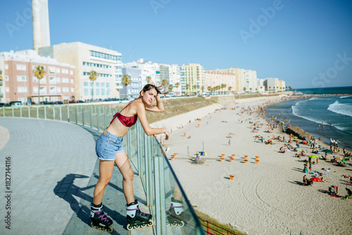 Woman wearing inline skates leaning on railing looking at beach on sunny day