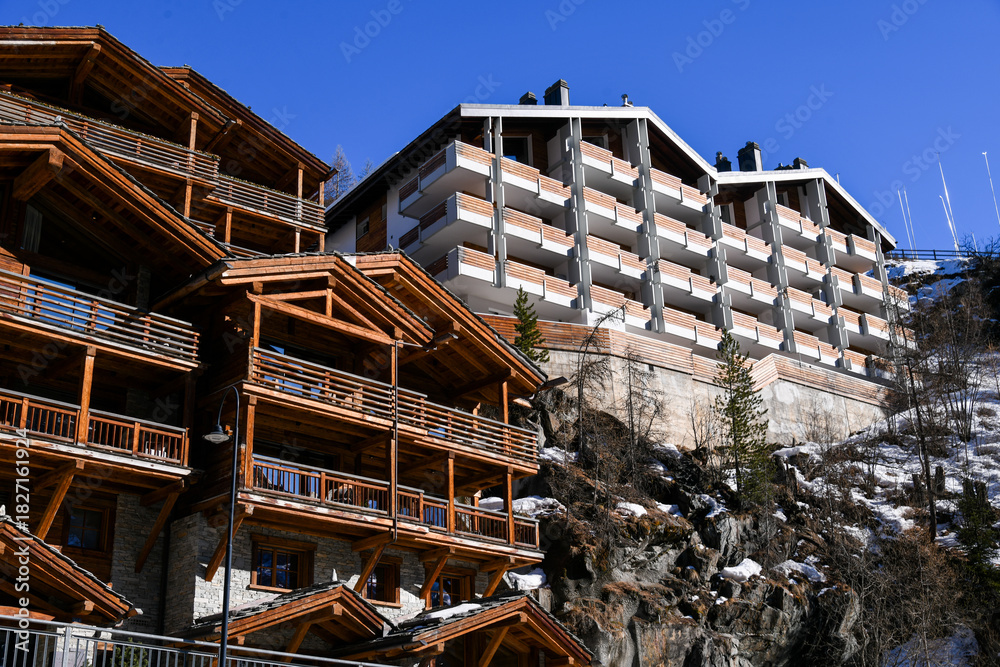 Obraz premium Traditional wooden alpine chalets and a modern mountain lodge located on a steep slope in Zermatt, Switzerland. Clear blue sky and winter daylight atmosphere.