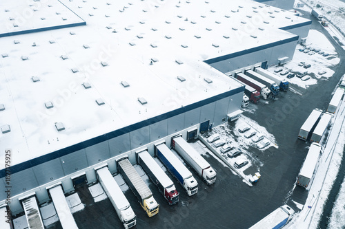 Winter aerial view of a large logistics warehouse in an industrial area with cars and trucks waiting to be unloaded and loaded.