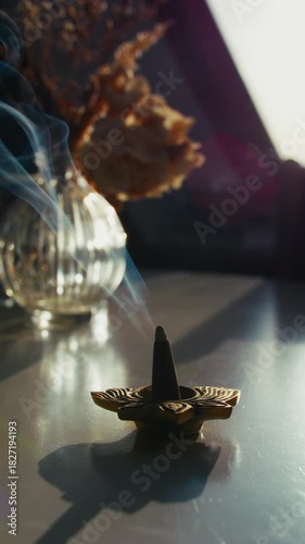 9x16 vertical close-up of smoke rising from a burning incense cone by a sunlit window on an autumn morning, with flowers on the table creating a serene, meditative, calming mood.