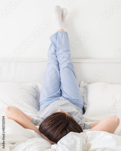 Fotografi Female legs raised up high and arms under her head lying on bed in bedroom weari